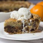 side view of slice of chocolate chip pie, ice cream on top dripping down the side of the pie on a white plate orange in background