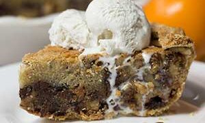 side view of slice of chocolate chip pie, ice cream on top dripping down the side of the pie on a white plate orange in background