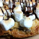 close up of baked magic cookie bar pizza shows toasted marshmallows, chocolate sauce on the cookie bar base