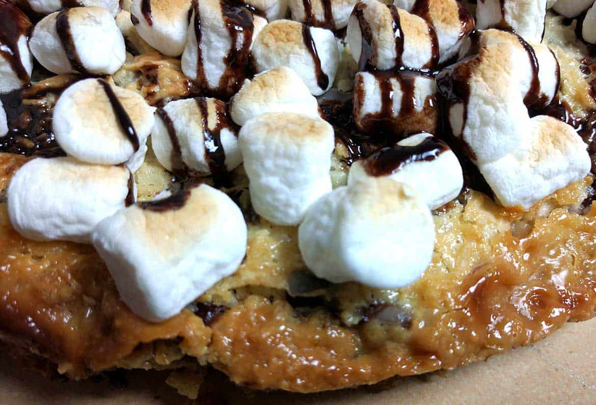 close up of baked magic cookie bar pizza shows toasted marshmallows, chocolate sauce on the cookie bar base