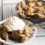 serving of baked apple cobbler with whipped cream on a blue and white plate with antique silver spoon and skillet of apple pandowdy in the background