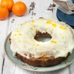 orange cranberry bundt cake with thick orange cranberry glaze orange zest on top blue plates and silver forks in right background three oranges in left background on white weathered wood table