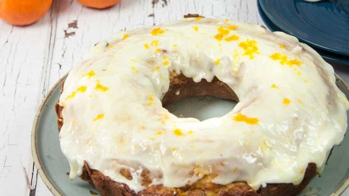 orange cranberry bundt cake with thick orange cranberry glaze orange zest on top blue plates and silver forks in right background three oranges in left background on white weathered wood table
