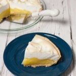 slice of lemon pie on blue plate with clear glass pie plate, pie server and lemon meringue pie with slice removed in background.