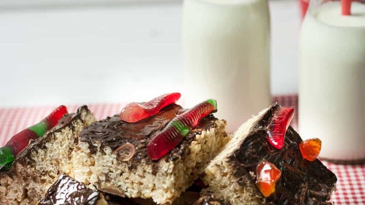 gummy worm treats with chocolate icing topped with gummy worms on a white plate on top of red white checked cloth