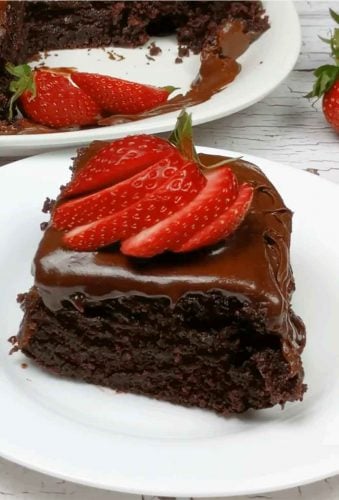 Slice of chocolate cake with sliced strawberries on top shown on white plate with whole cake in background