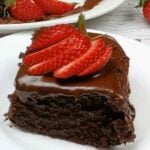 slice of chocolate cake on a white plate with sliced strawberries on top of chocolate frosting with whole cake in background