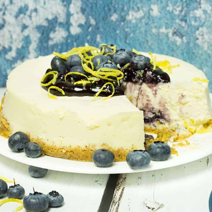 lemon blueberry cheesecake on white plate with blueberries and lemon peel piled on top, blueberries on the side, and a slice removed. shows a blue background on top of a white wooden table