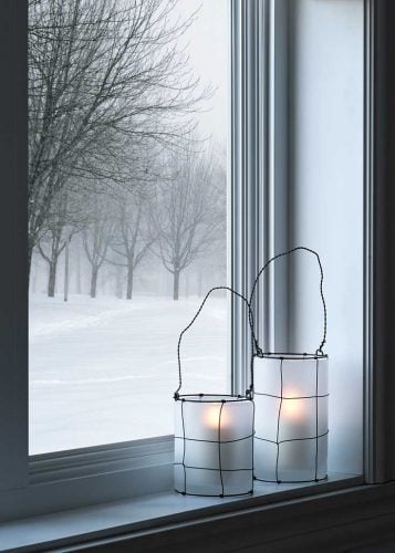 winter scene looking out a window you can see a snowstorm with two white candle holders, lit candles inside, and wrapped in wire with wire handles on the window sill. 