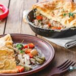 recipe image of puff pastry topped Chicken Pot Pie on a brown plate with brown fork, shows cast iron skillet in background with serving removed, and red chili pepper to the left