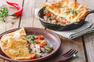 recipe image of puff pastry topped Chicken Pot Pie on a brown plate with brown fork, shows cast iron skillet in background with serving removed, and red chili pepper to the left