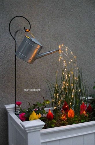 metal watering can on a black iron hook staked into a flower box with fairy lights coming out of the spout
