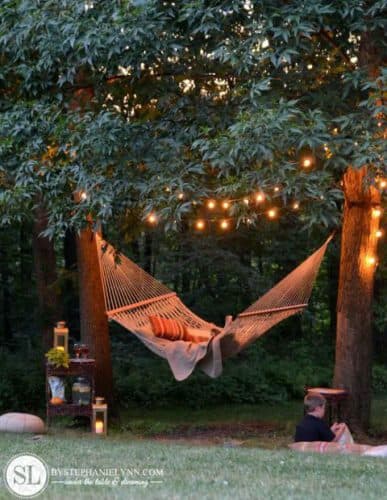 canvas hammock with orange pillow between two posts with tree branches overhead filled with fairy lights