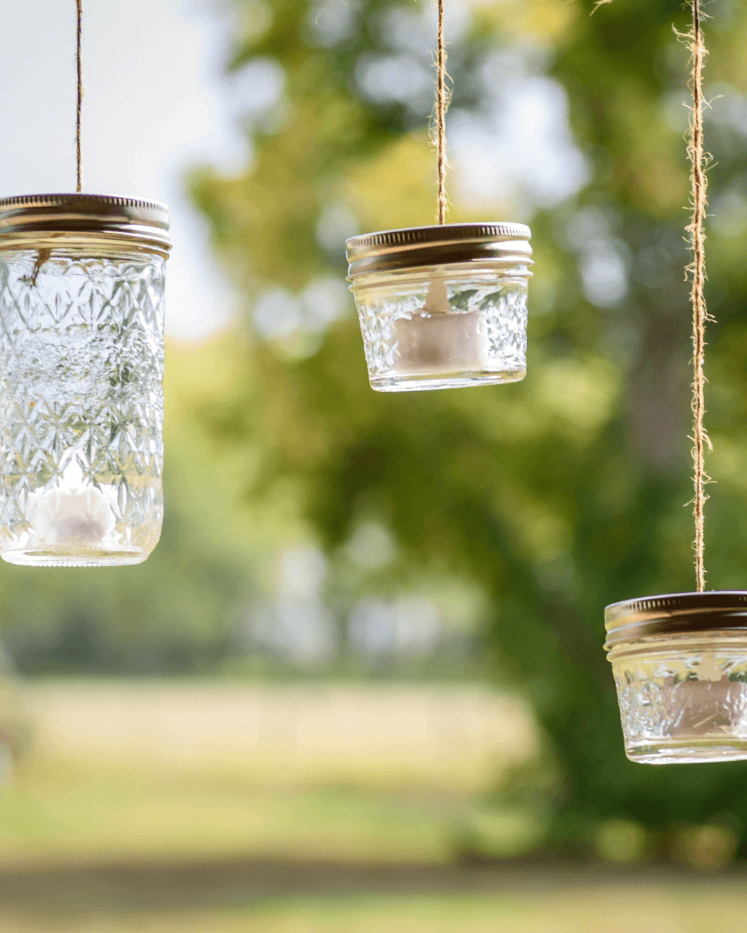 Three glass jars with metal lids and candles inside hang outdoors from twine, creating a magical touch reminiscent of DIY fairy light projects, set against a blurred background of trees and greenery.