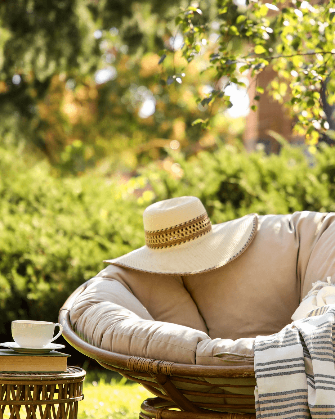 Round cushioned wicker chair with a straw hat rests outdoors beside a small table with a cup and saucer; DIY fairy light projects twinkle nearby, adding charm amid lush greenery and sunlight in the background.