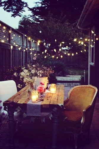 wooden backyard table with white chair and gold coloured chair, flowers and candles on table with patio lights strung from fence to back of house