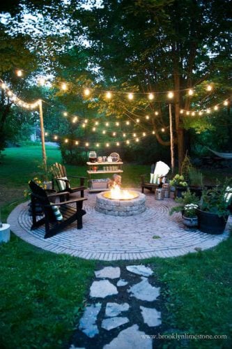 backyard fire pit with chairs place around it, stone path leads to conversation area, and several strands of string lights across entire area