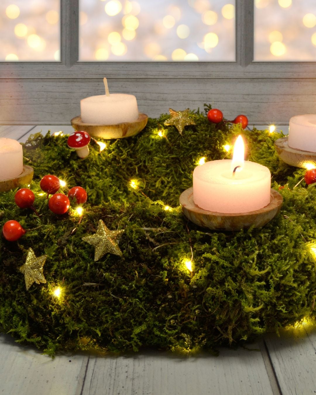 A moss-covered wreath decorated with red berries, gold stars, small wooden mushrooms, string lights from DIY fairy light projects, and four tealight candles—one glowing—rests on a wooden surface.