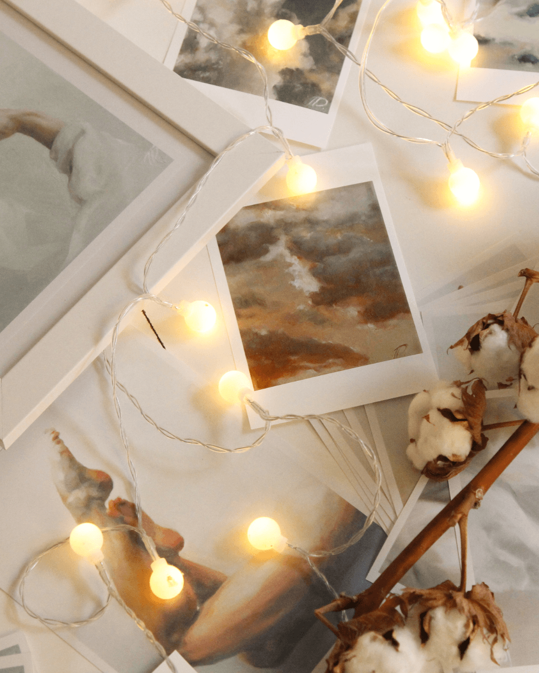 A string of warm fairy lights is draped over art prints and cotton stems on a flat surface, perfect inspiration for DIY fairy light projects.