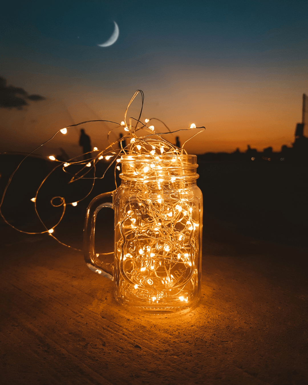 A mason jar filled with string lights sits on a surface outdoors at sunset, featuring a crescent moon and a silhouette of a person in the background—perfect inspiration for DIY fairy light projects.