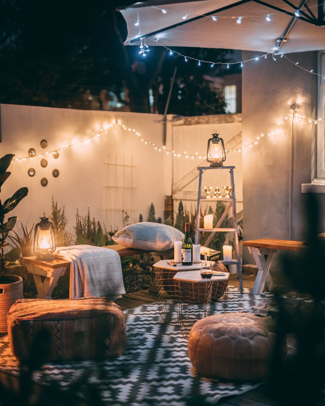 Cozy outdoor patio at night with string lights, lanterns, pillows, a rug, benches, and a small table with wine and glasses. DIY fairy light projects and decorative plants surround the inviting seating area.
