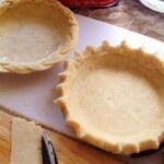 2 unbaked pie crust shells on white and black cutting board and red rolling pin on the side