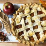 lattice topped apple pie with measuring spoons, cinnamon sticks, apples on a wooden board