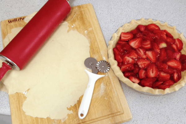 A rolling pin, pastry cutter, and rolled-out dough rest on a cutting board beside an unbaked strawberry pie with a fluted crust, ready to be filled with delicious strawberry pie filling.