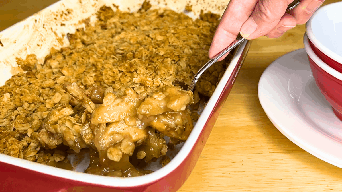 A hand scoops apple crisp from a red baking dish with a spoon. Showcasing an easy apple crisp recipe, the dessert has a golden, crumbly topping and is served near a stack of red and white bowls.