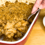 A hand scoops apple crisp from a red baking dish with a spoon. Showcasing an easy apple crisp recipe, the dessert has a golden, crumbly topping and is served near a stack of red and white bowls.