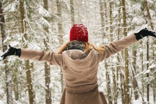 woman with camel coloured coat and red knitted hat outdoors with arms outstretched in front on snow covered trees