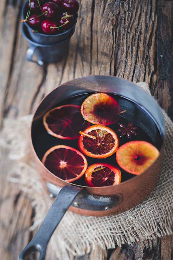 copper pot on wooden table and burlap cloth under pot with mulled wine, slices of oranges and spices floating on top.