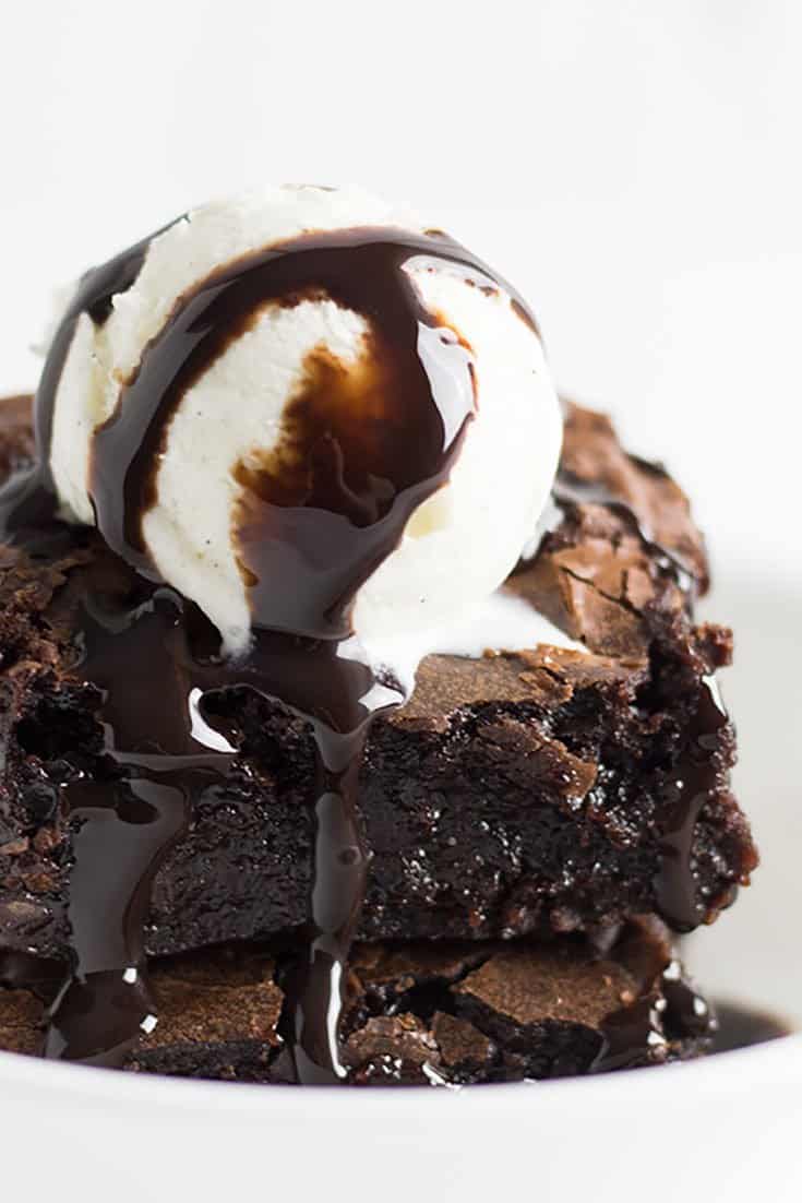 generous piece of chocolate brownie with scoop of vanilla ice cream chocolate sauce running over everything in a white bowl