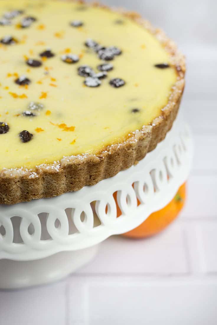 pale yellow custard with chocolate chips filling topped with orange zest tart on a white cake plate with scalloped edging and orange underneath