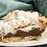 slice of chocolate pudding pie on a white china plate topped with whipping cream and multi coloured sprinkles. shows two blue coffee mugs and blue napkins in background