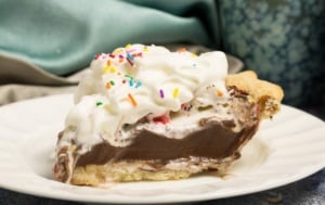 slice of chocolate pudding pie on a white china plate topped with whipping cream and multi coloured sprinkles. shows two blue coffee mugs and blue napkins in background