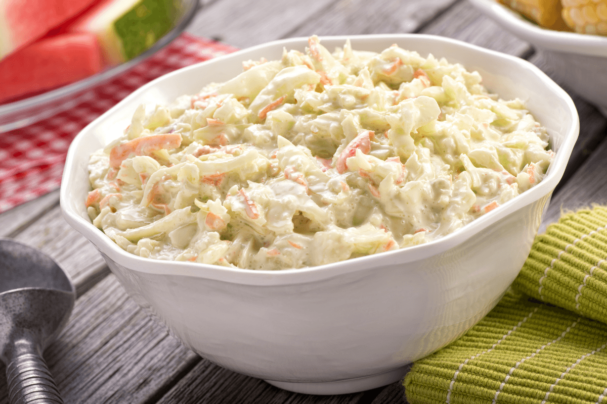 A white bowl filled with creamy coleslawโperfect for any easy BBQ party menuโsits on a wooden table next to a green striped cloth and a metal serving spoon.