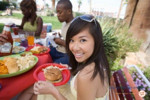girl with long black hair and sunglasses on her head with a red picnic plate and hamburger and salad on it, with friends eating outside in the backgroun