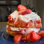 A homemade strawberry shortcake recipe comes to life in this delightful dessert, featuring layers of biscuit, whipped cream, and sliced strawberries on a blue plate, with extra strawberries nestled around the base.