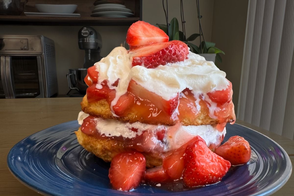 A homemade strawberry shortcake recipe comes to life in this delightful dessert, featuring layers of biscuit, whipped cream, and sliced strawberries on a blue plate, with extra strawberries nestled around the base.