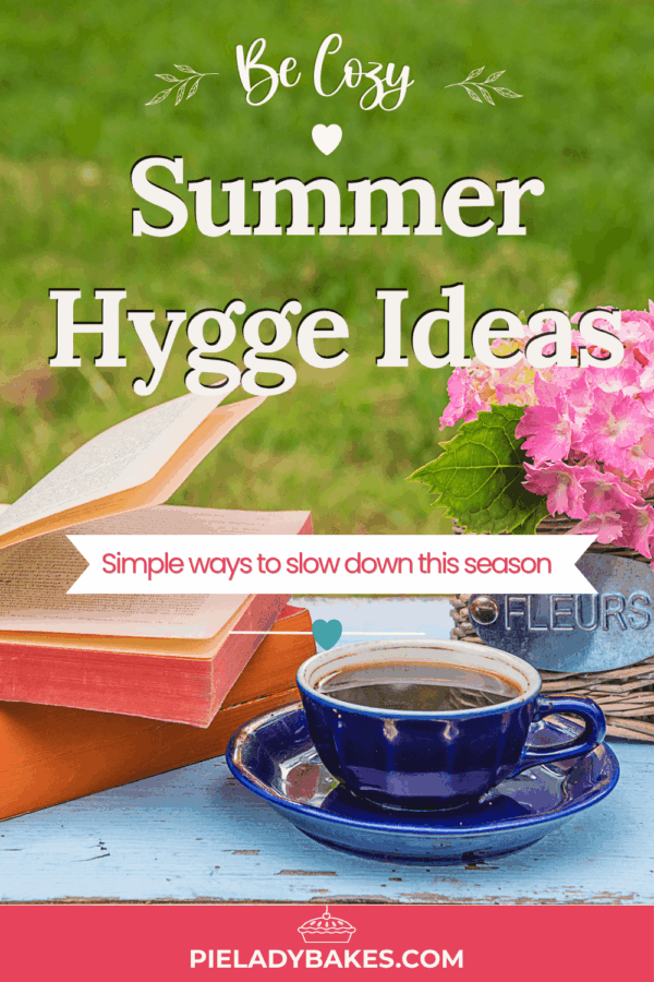 A blue cup of coffee, an open book, and pink flowers sit on a blue table outdoors, with text sharing how to hygge and inspiring summer hygge ideas from pieladybakes.com.