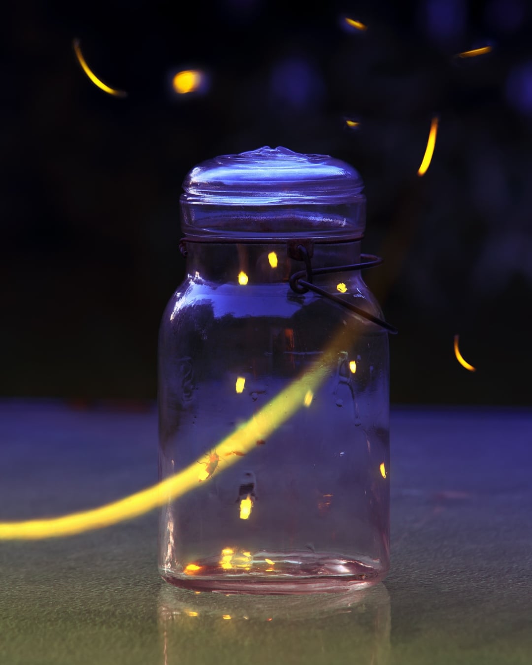 A glass jar with fireflies inside and a few glowing fireflies flying around it, set on an outdoor surface at dusk—capturing the magic of summer hygge and simple joys that can improve your daily life.