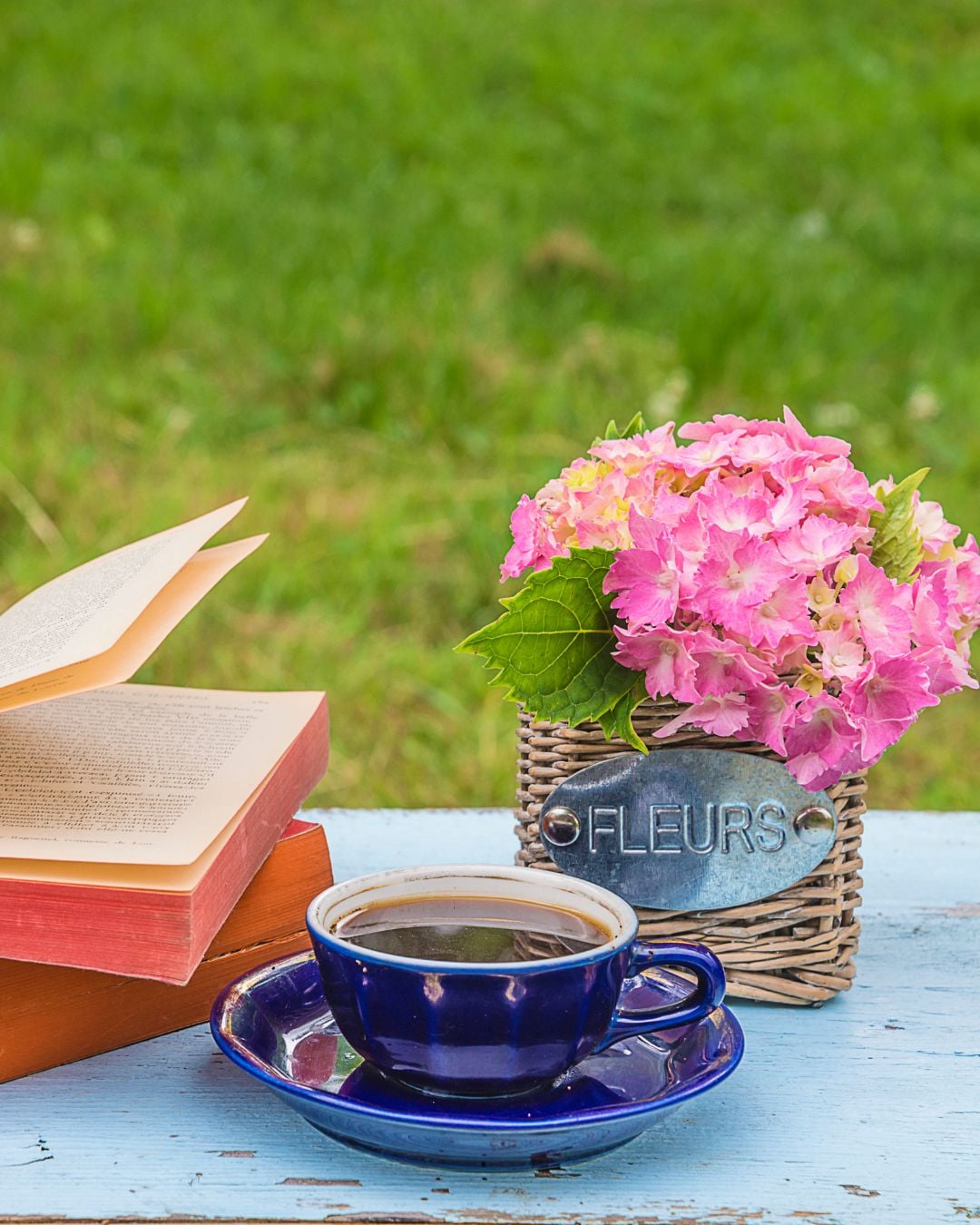 A blue cup of coffee, an open book, and a pink hydrangea bouquet in a "Fleurs" basket sit on a blue wooden table outdoors with green grass—perfect summer hygge for relaxing moments.