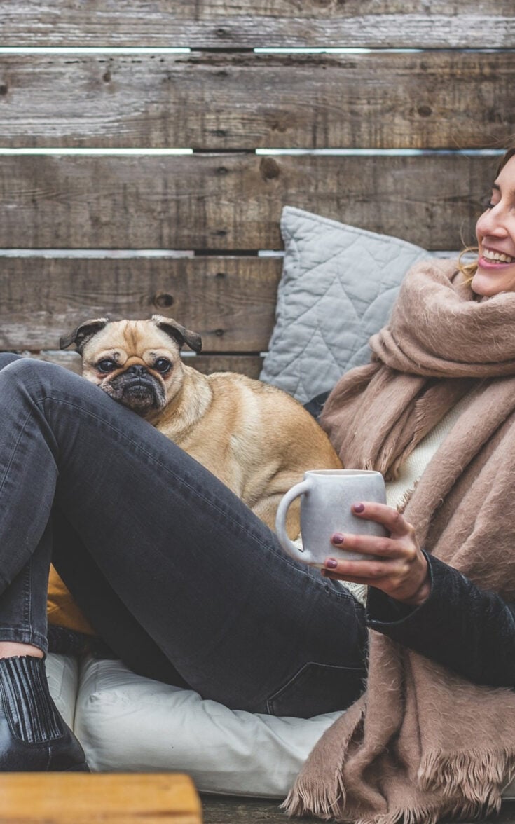 woman with pink scarf and small dog on her lap holding a white mug