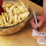 A person writes “Sept 8” on a freezer bag with a marker next to a bowl filled with sliced apples coated in cinnamon, prepping apple pie filling before freezing.