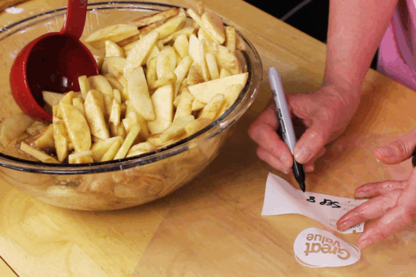 A person writes โSept 8โ on a freezer bag with a marker next to a bowl filled with sliced apples coated in cinnamon, prepping apple pie filling before freezing.