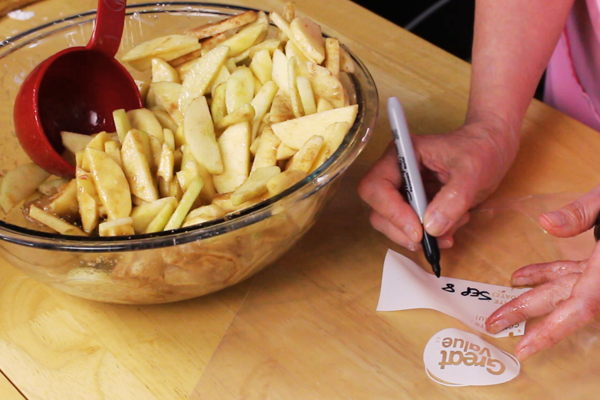A person writes “Sept 8” on a freezer bag with a marker next to a bowl filled with sliced apples coated in cinnamon, prepping apple pie filling before freezing.