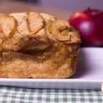 apple pie bread side view shows cinnamon, butter, apples, brown sugar on a white plate green & white checked cloth and red apple in background