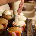 woman's hands piping cream cheese frosting onto several cupcakes and shows plain cupcakes too