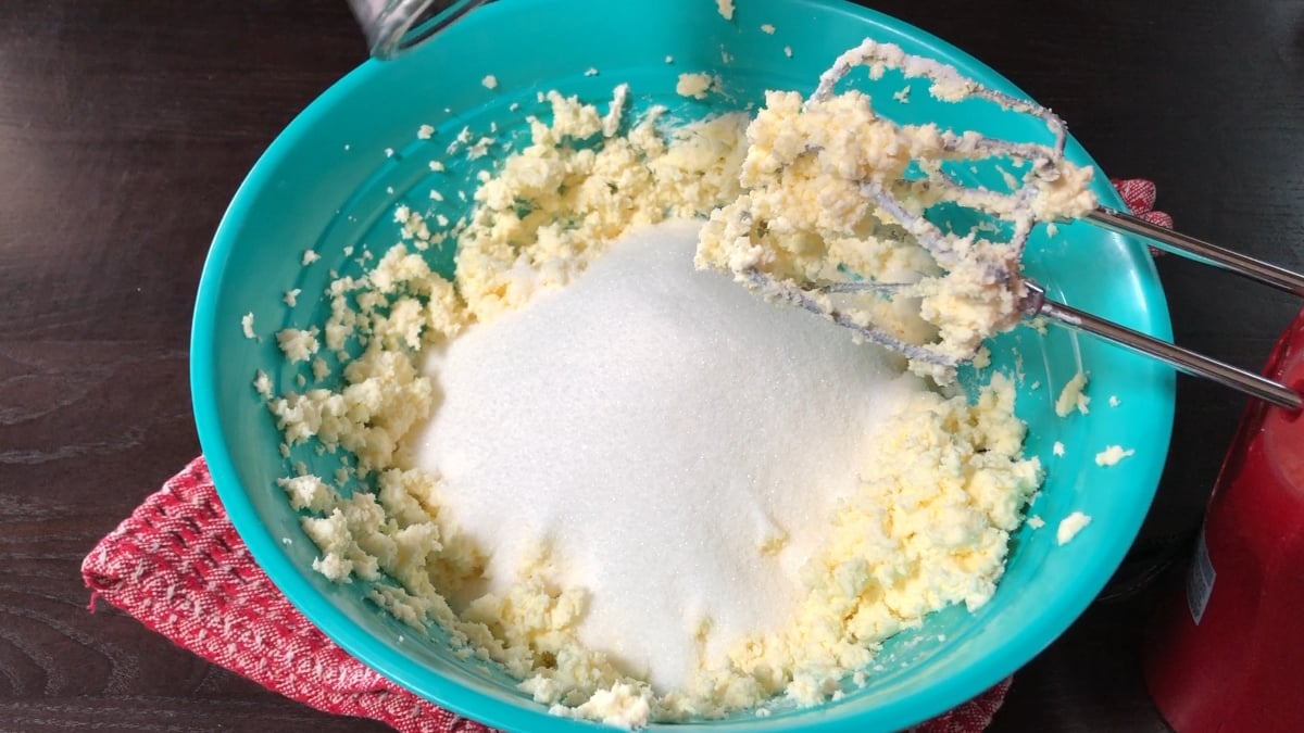 A blue bowl with creamed butter and sugar is mixed for an easy strawberry pound cake using an electric hand mixer, all set on a red cloth atop a dark surface.
